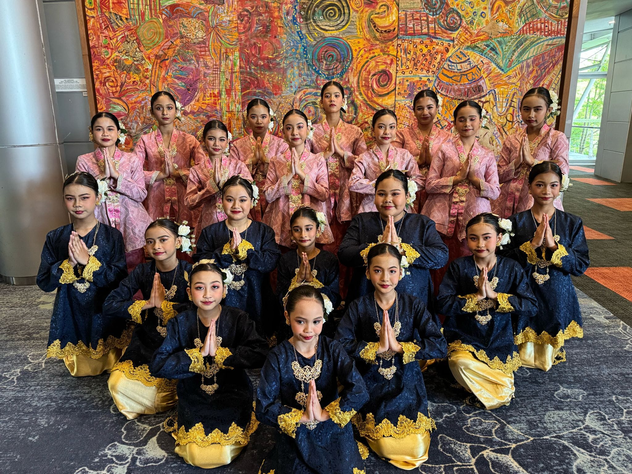 A Malay dance group poses in a triangular formation, smiling confidently in traditional costumes before their performance.